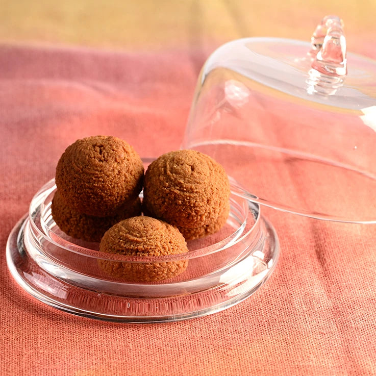 LEGÙ Pasticcini morbidi alla mandorla: 4 dolcetti senza glutine e ricchi di proteine vegetali, su alzata in vetro con cupola, su telo arancione.