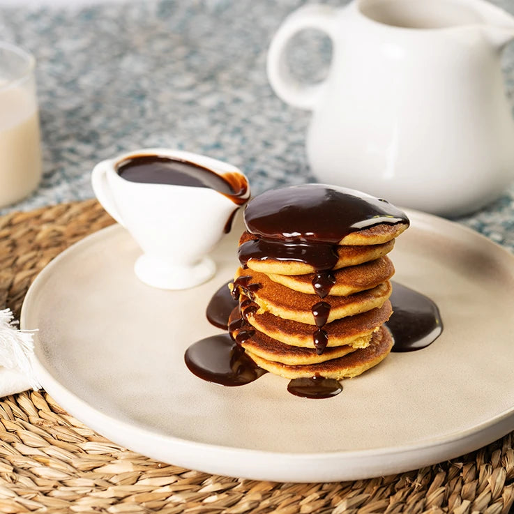 Stack di pancakes LEGÙ al cioccolato, soffici e dorati, su piatto in ceramica con salsa al cioccolato e bricco di latte. Preparato per Pancakes Biologico, senza glutine.